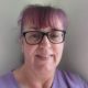 A woman with light skin, purple hair and bangs, wearing black glasses and a purple shirt, smiles at the camera while standing against a light-colored wall.