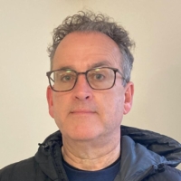 A middle-aged man with short, curly grey hair and glasses is wearing a dark jacket and looking straight at the camera in front of a plain, light-coloured background.