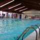 Indoor swimming pool with several swimmers in the water. The pool area has a tiled deck, metal ladders, and equipment like lifebuoys and kickboards along the walls. Ceiling beams and lights are visible above.