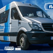 A silver Flexibus minibus is parked on a road under a blue sky. The vehicle features blue stripes and the Flexibus logo. App store icons and partner logos appear in the corners of the image.