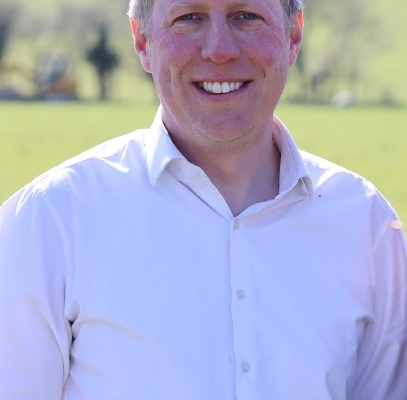 A man with short light brown hair, wearing a white button-up shirt, stands outside smiling with green fields and trees blurred in the background.