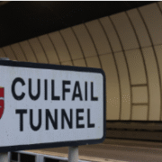 A sign with a red crest reads "Cuilfail Tunnel" in front of the entrance to a modern, curved tunnel with white tiled walls and dim lighting.