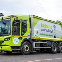 A green and yellow garbage truck parked on a street, featuring the slogan "Rethink your rubbish. Reduce, Reuse, Recycle" on its side, near a metal fence on a cloudy day.