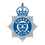 The Sussex Police logo, featuring a blue and silver eight-pointed star with a crown on top, and a circular emblem in the center displaying "Sussex Police" around a shield with six birds and a cross.