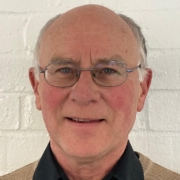 A smiling older man with glasses, short gray hair, and a beige sweater over a black collared shirt, standing in front of a white brick wall.