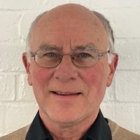 A smiling older man with glasses, short gray hair, and a beige sweater over a black collared shirt, standing in front of a white brick wall.