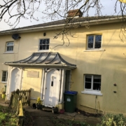 A cream-coloured, two-storey house with a unique sloped metal canopy over two white front doors, surrounded by a garden, trees, and a green bin near the entrance on a sunny day.
