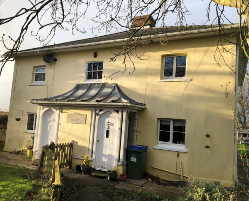 A cream-coloured, two-storey house with a unique sloped metal canopy over two white front doors, surrounded by a garden, trees, and a green bin near the entrance on a sunny day.