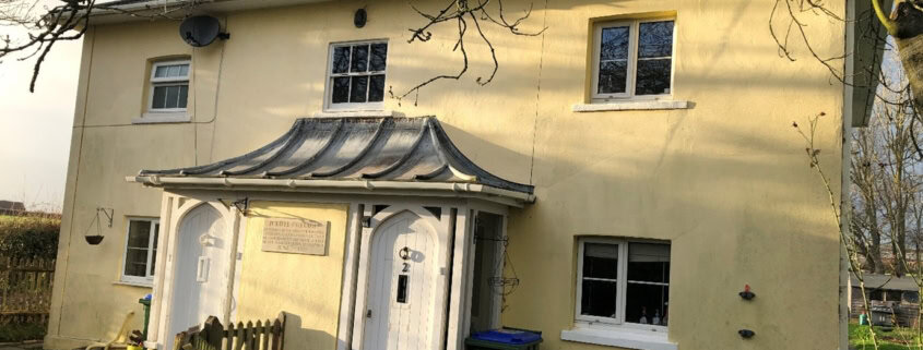 A cream-coloured, two-storey house with a unique sloped metal canopy over two white front doors, surrounded by a garden, trees, and a green bin near the entrance on a sunny day.