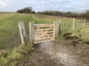A wooden gate stands open on a muddy path through grassy fields, bordered by wire fencing and weathered wooden posts, under a mostly cloudy sky with patches of blue.