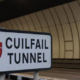 A sign reading "Cuilfail Tunnel" with a red shield emblem, positioned in front of the entrance to a modern, curved tunnel.