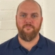 A man with a shaven head and a brown beard wearing a navy blue polo shirt stands in front of a white brick wall, looking directly at the camera with a neutral expression.