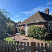 A brick bungalow with a paved patio, outdoor seating, and a wooden fence in the foreground.