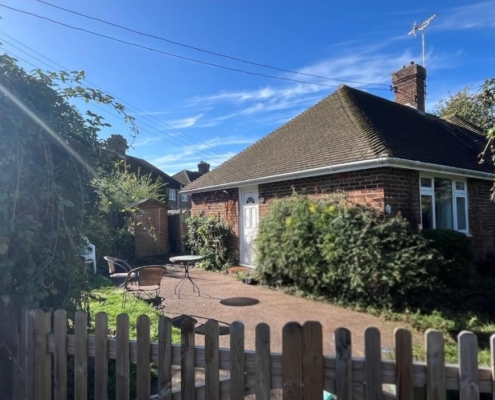 A brick bungalow with a paved patio, outdoor seating, and a wooden fence in the foreground.