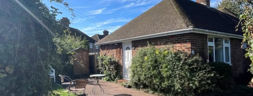 A brick bungalow with a paved patio, outdoor seating, and a wooden fence in the foreground.